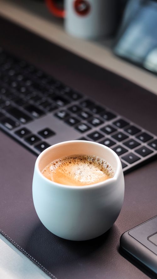 Free A fresh cup of coffee sits on a desk with a keyboard in a modern workspace. Stock Photo