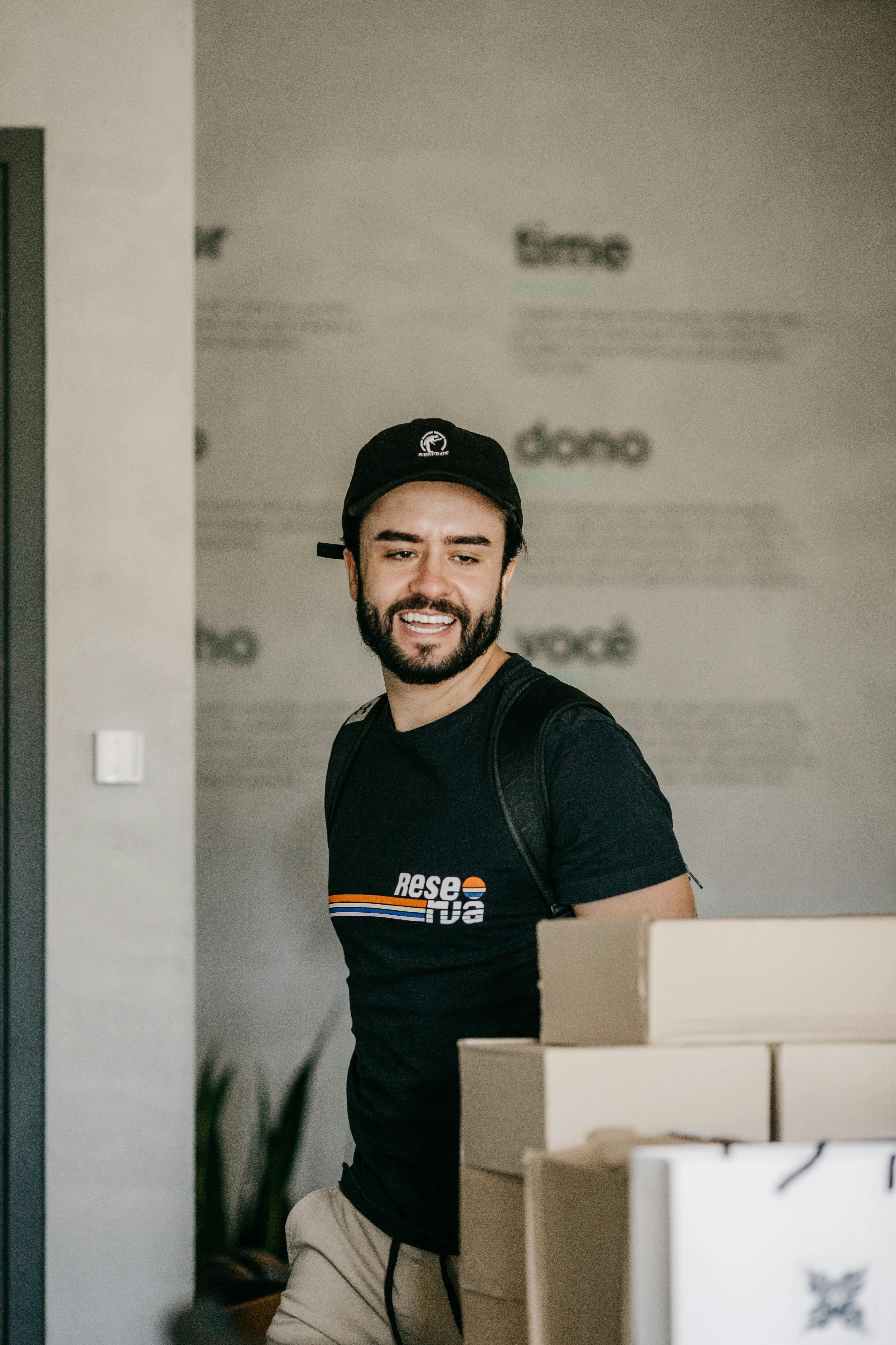 Casual man with beard wearing cap and t-shirt in a modern office, surrounded by boxes.