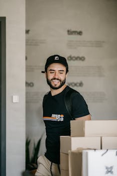 Casual man with beard wearing cap and t-shirt in a modern office, surrounded by boxes.