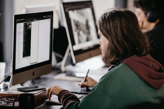 Person in a hoodie working at a desktop computer in an office setting, focused and engaged.