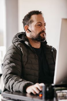 Focused man wearing a jacket and headset working at a computer indoors.