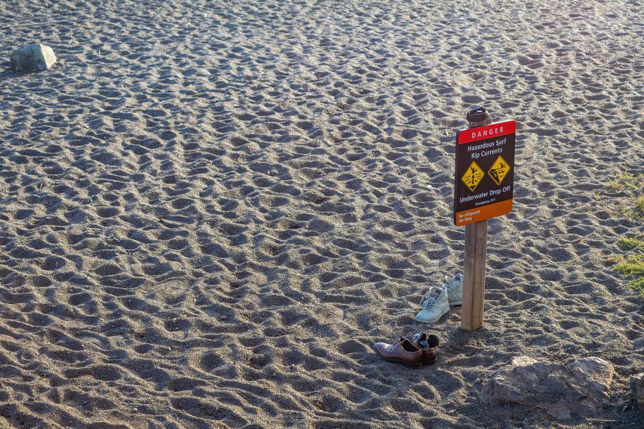 A beach warning sign stands on the sandy shore with two pairs of shoes nearby, emphasizing caution.