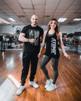 Fit couple smiling and posing in a bright, modern gym.