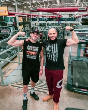 Two men posing and flexing their biceps in a lively gym setting, showcasing fitness together.