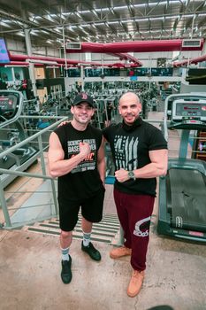 Two men posing and smiling in a well-equipped gym, showcasing fitness camaraderie.