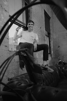A black and white portrait of a man smiling while sitting casually on a sofa by an industrial window.