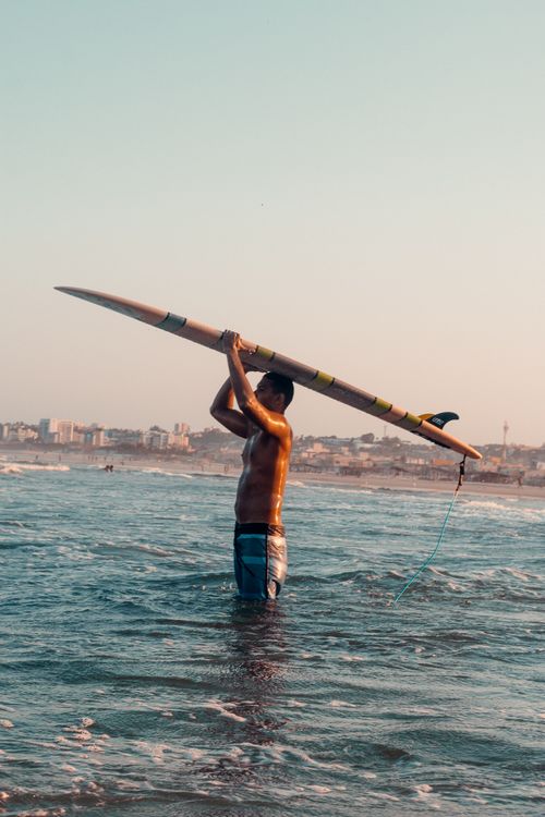 Free Man standing in ocean with surfboard overhead at sunset, preparing to surf. Stock Photo