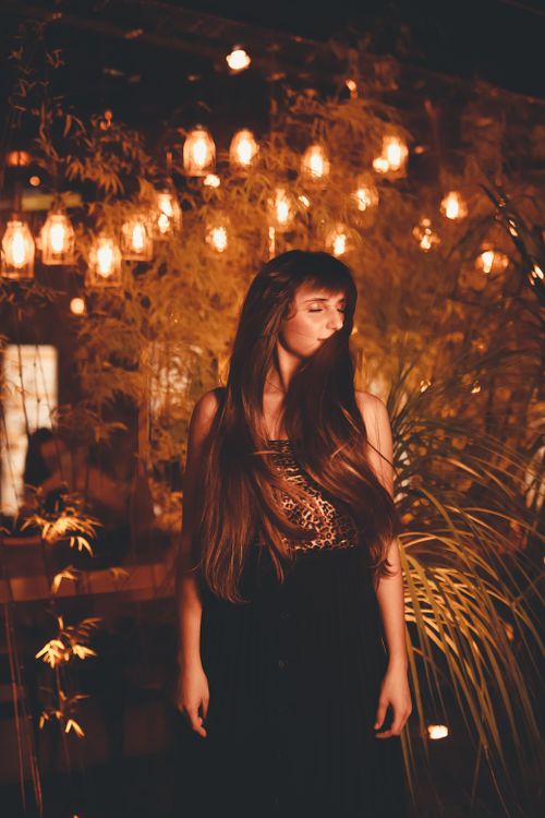 Free A woman with long hair poses gracefully outdoors in a garden illuminated by warm lights, creating a serene night atmosphere. Stock Photo