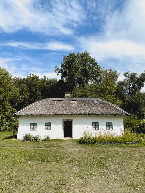 Free Quaint countryside cottage with thatched roof surrounded by lush greenery under blue sky. Stock Photo
