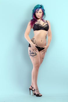 A young woman with dyed hair and tattoos poses in stylish lingerie against a blue background.