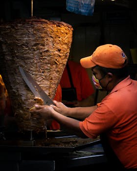 A man slices seasoned al pastor meat from a vertical spit in a busy food stall.