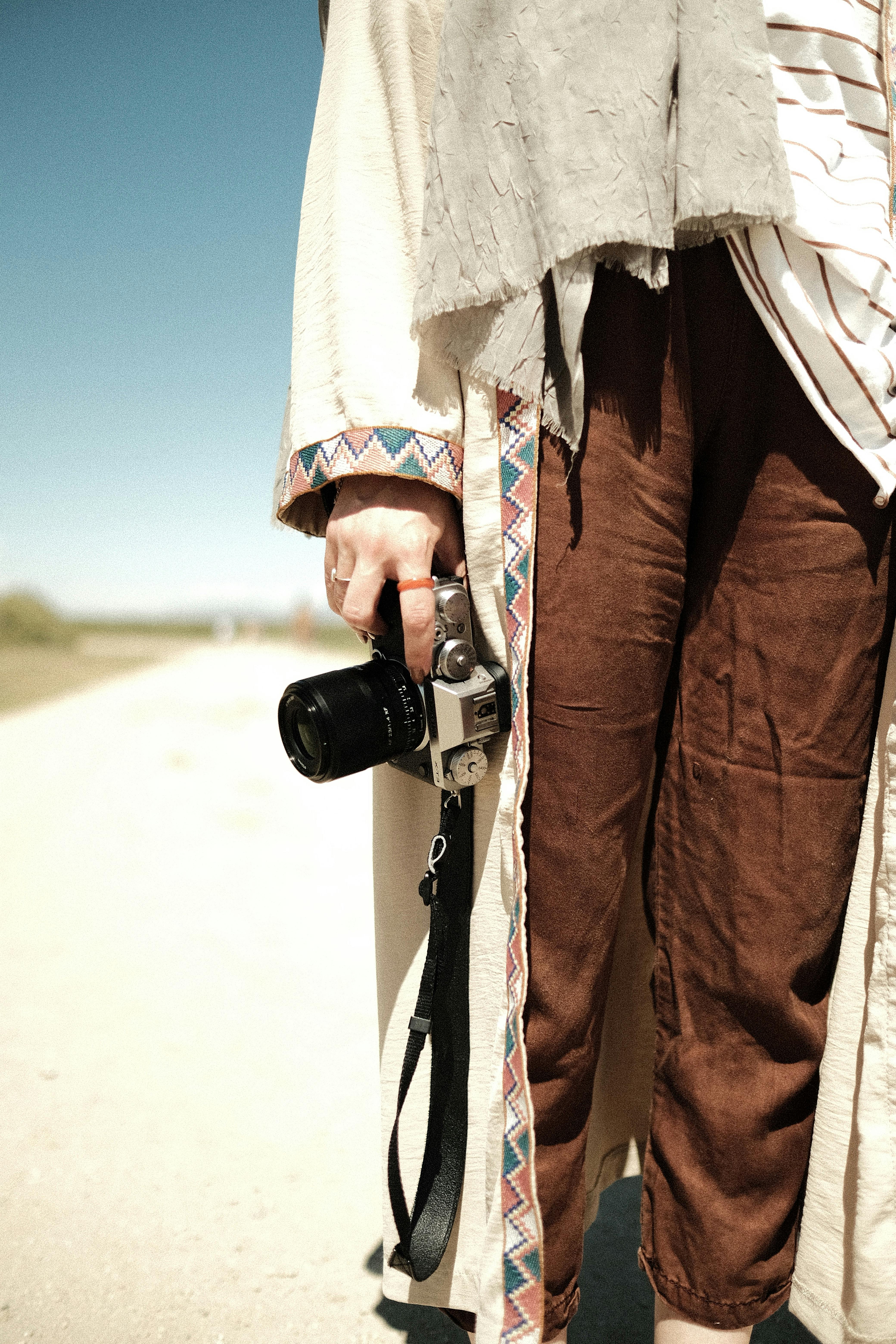 A fashionable individual holding a camera outdoors in stylish brown pants and a patterned coat.