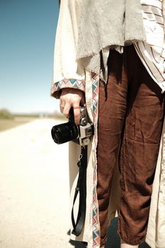 A fashionable individual holding a camera outdoors in stylish brown pants and a patterned coat.