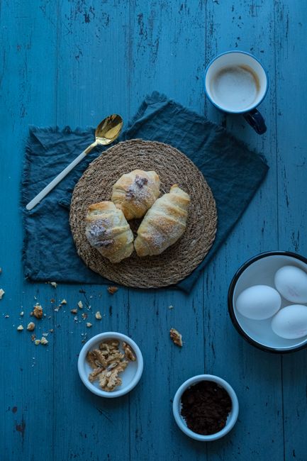 Flat lay of croissants, coffee, and ingredients on a rustic blue wooden table.