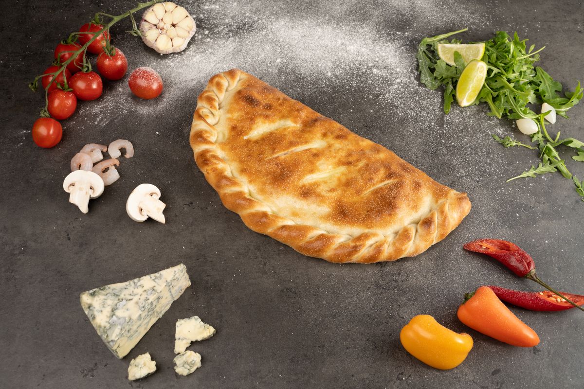 Golden half-moon calzone with crimped edge surrounded by mushrooms, blue cheese, tomatoes and peppers
