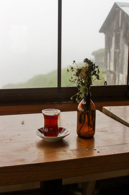 A glass of Turkish tea on a wooden table beside a vase of wildflowers near a misty window.