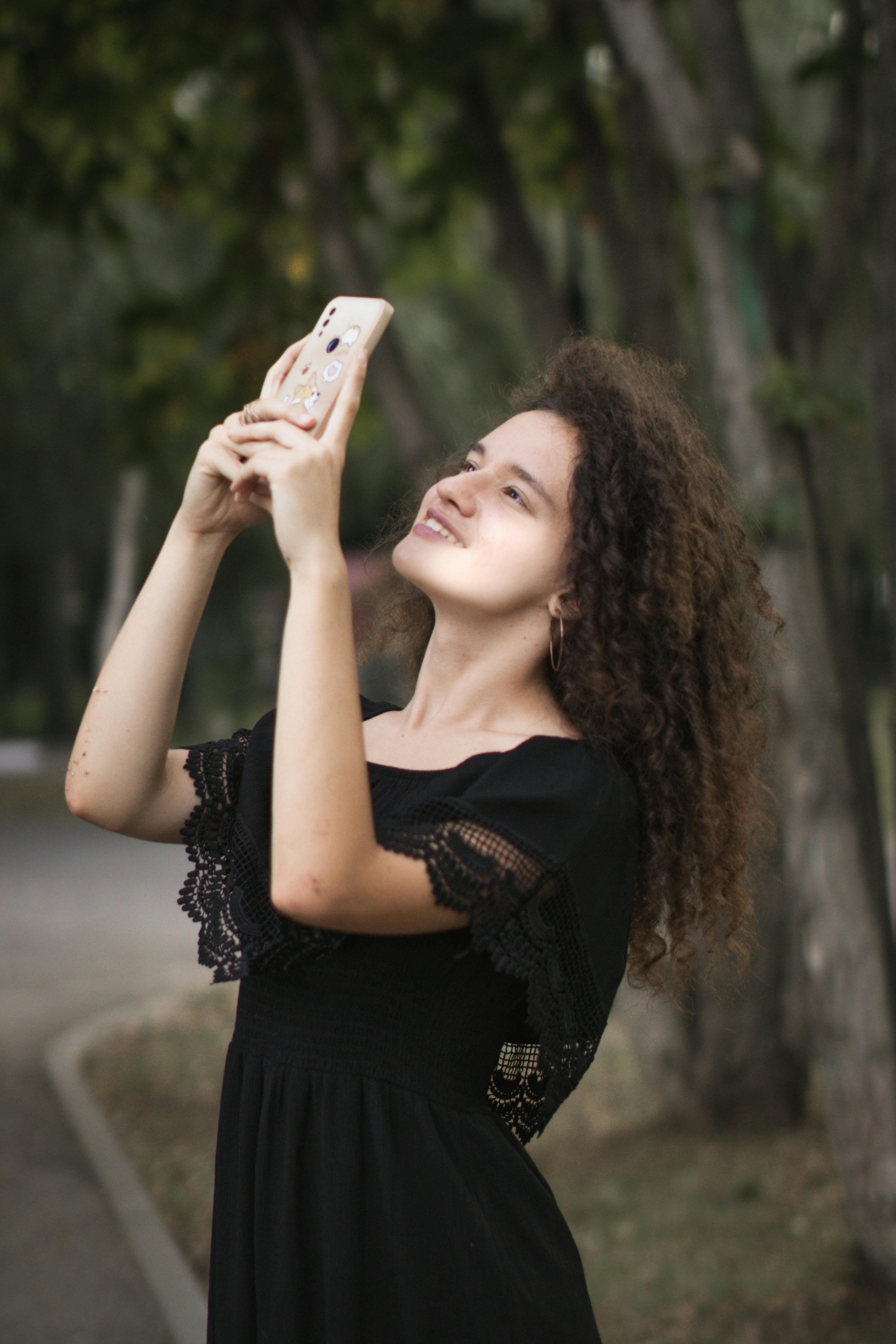Young woman in black dress with curly hair smiles while taking a photo outdoors.