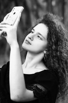 A black and white portrait of a young woman taking a selfie with a smartphone outdoors.