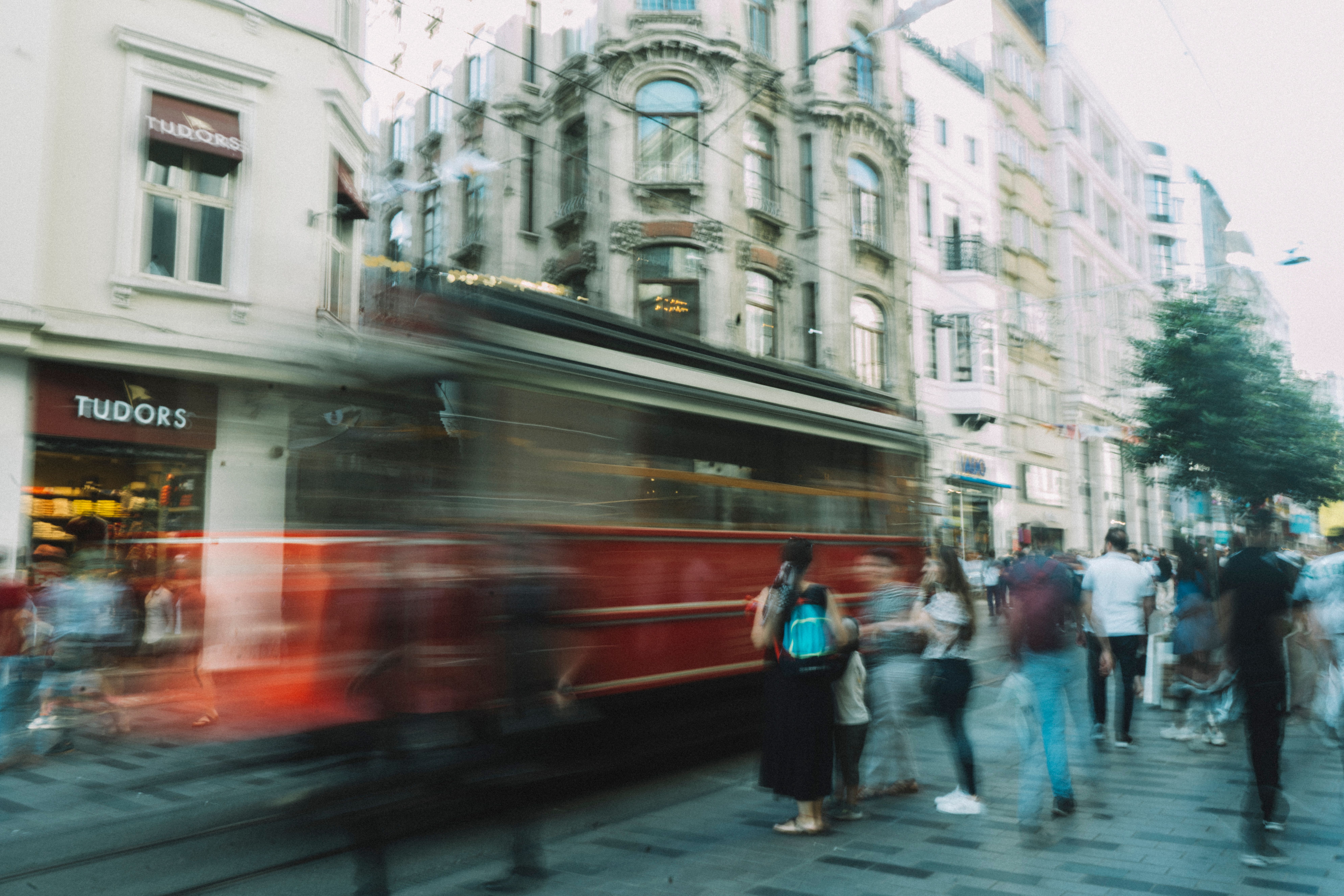 Cena de uma rua movimentada com um bonde clássico em movimento, cercada por edifícios históricos e pessoas caminhando, refletindo o cotidiano urbano e o transporte público.