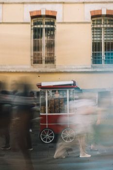 A dynamic urban scene capturing blurred motion of people around a vibrant red food cart against building facades.
