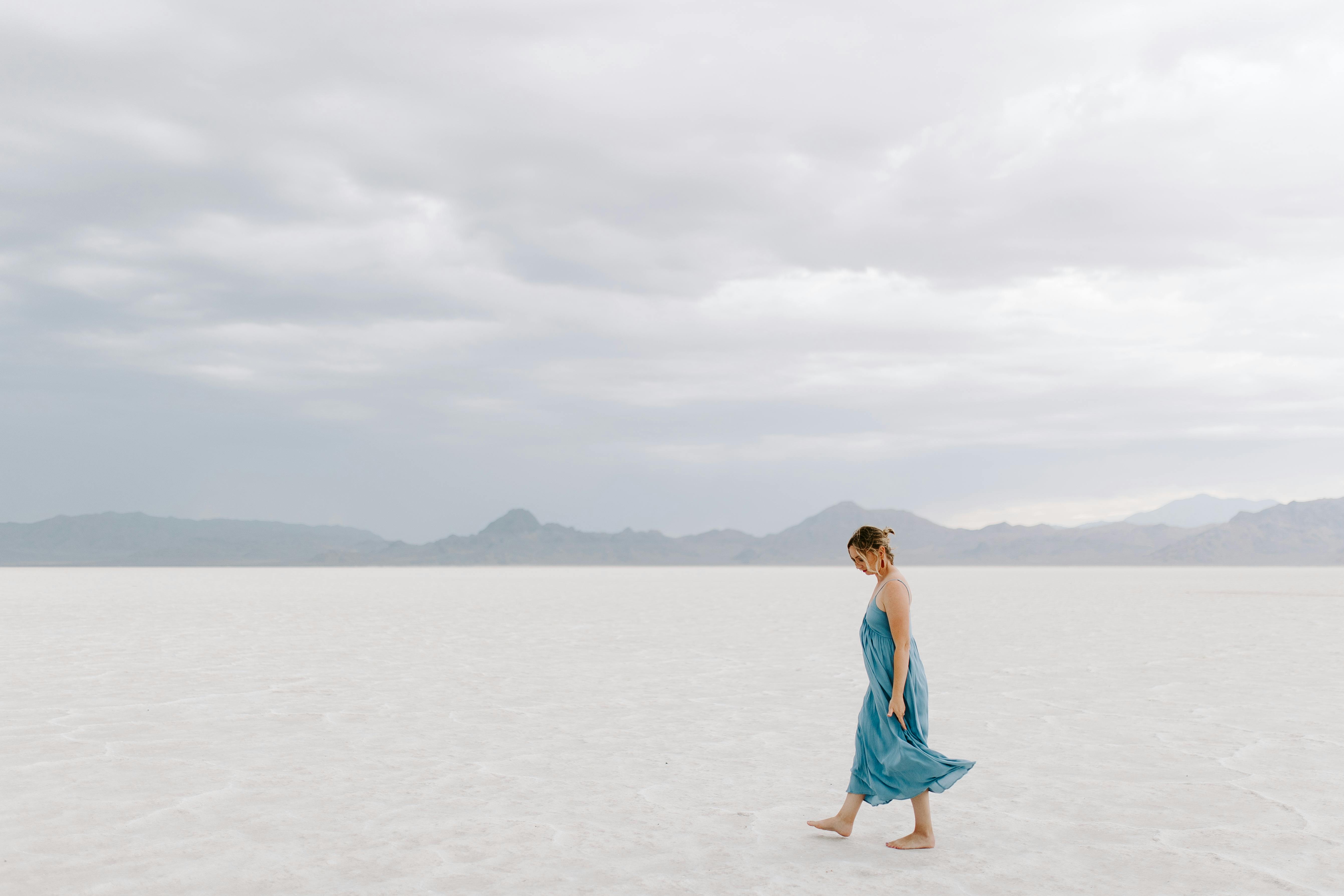 A woman in a blue dress walks alone on out vast white salt flats under a cloudy sky.