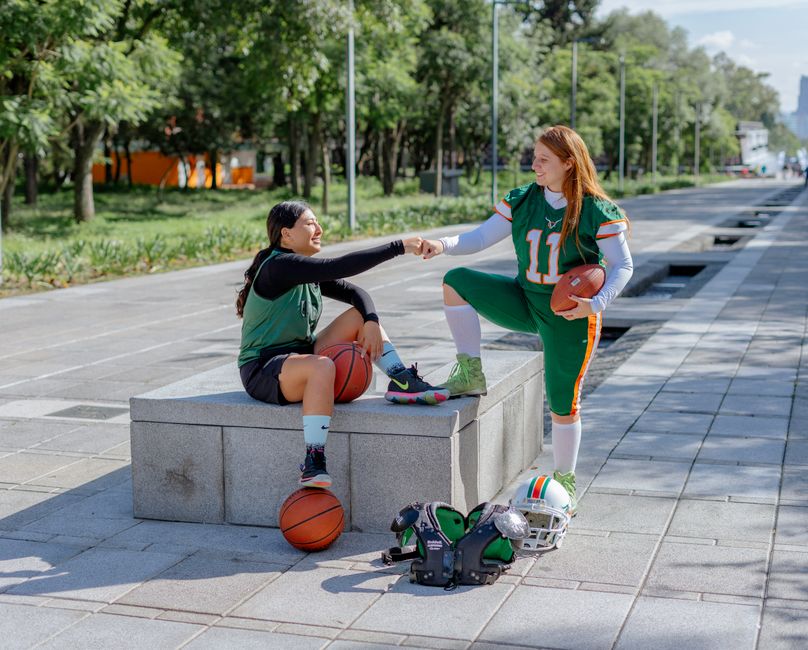 Two female athletes share a fist bump while sitting outdoors, holding sports equipment in Mexico City.