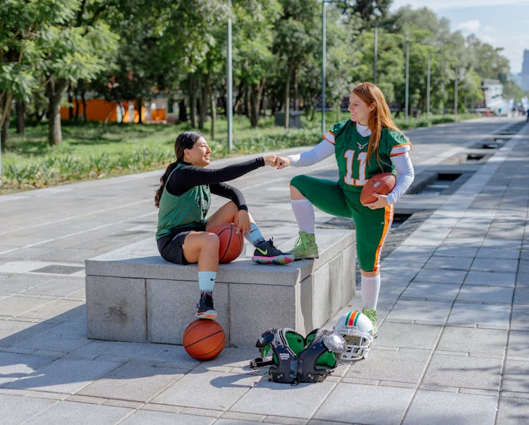 Girl In Basketball Clothing And Girl In American Football Clothing Fist Bumping 