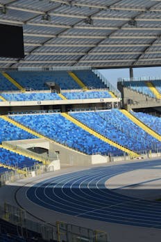 A spacious empty stadium with blue and yellow seats and an athletics track under a clear roof.