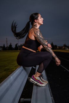 Tattooed woman in workout attire crouching on a bench during nighttime, exuding strength.