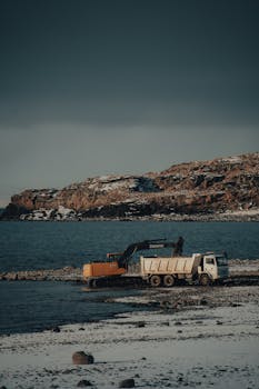 An excavator and truck work on a snowy coast with a rocky landscape in the background.