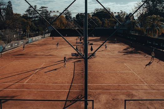 Tennis players compete on clay court with autumn foliage in the background.