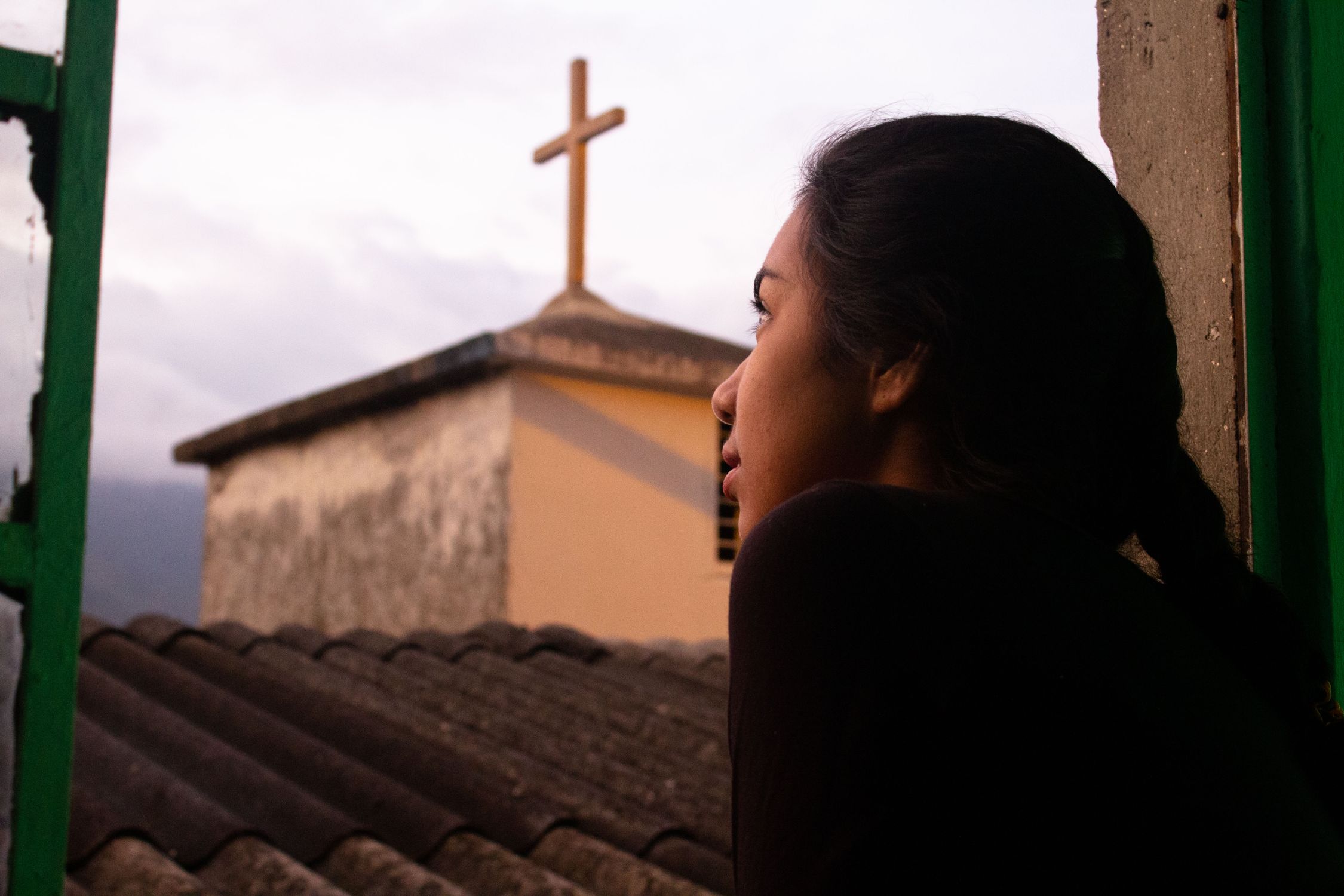 A woman looks out a window at a church cross during sunset in Medellín, Colombia.