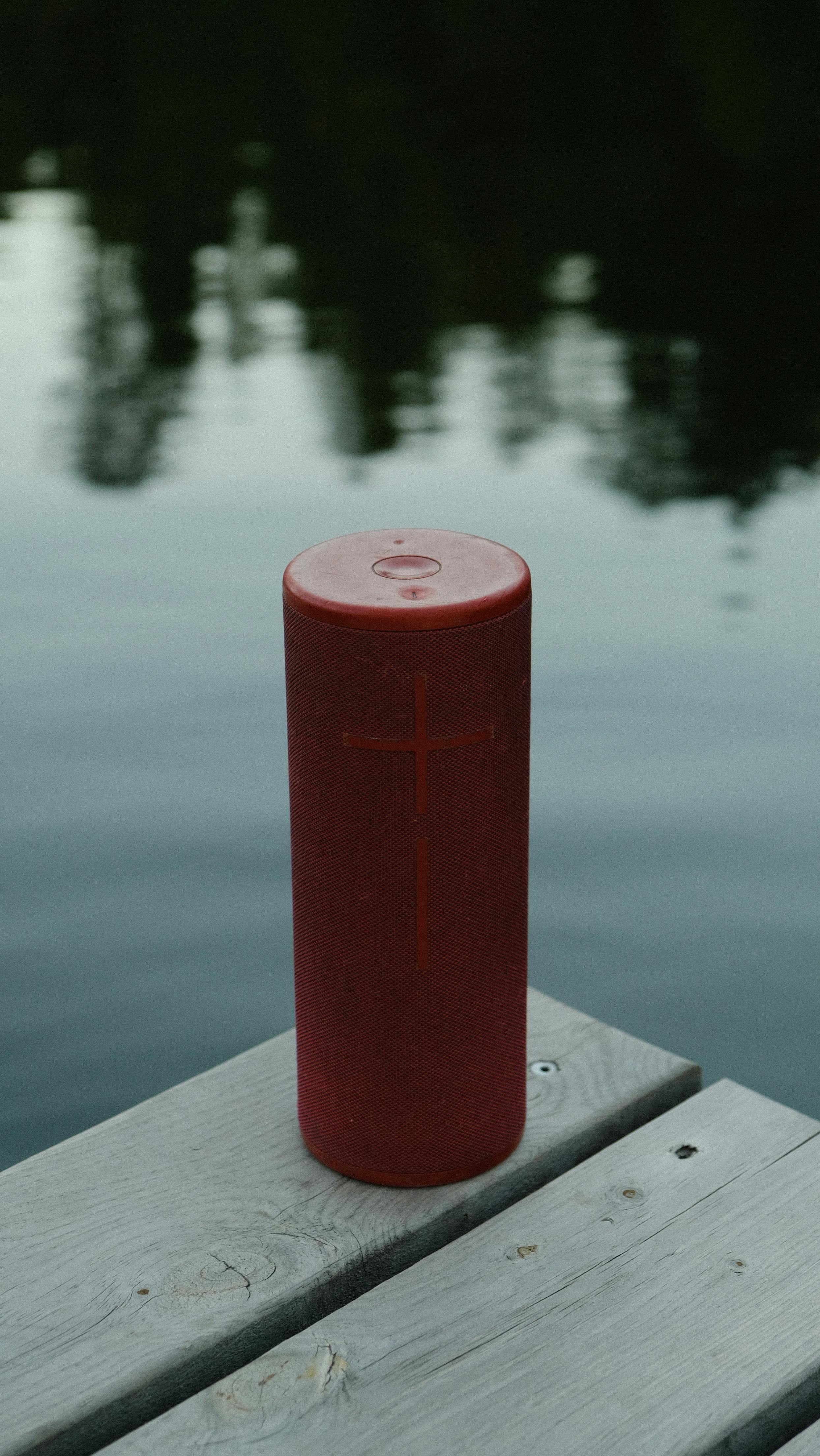 Red wireless speaker on a wooden dock by a serene lake. Ideal for outdoor music.