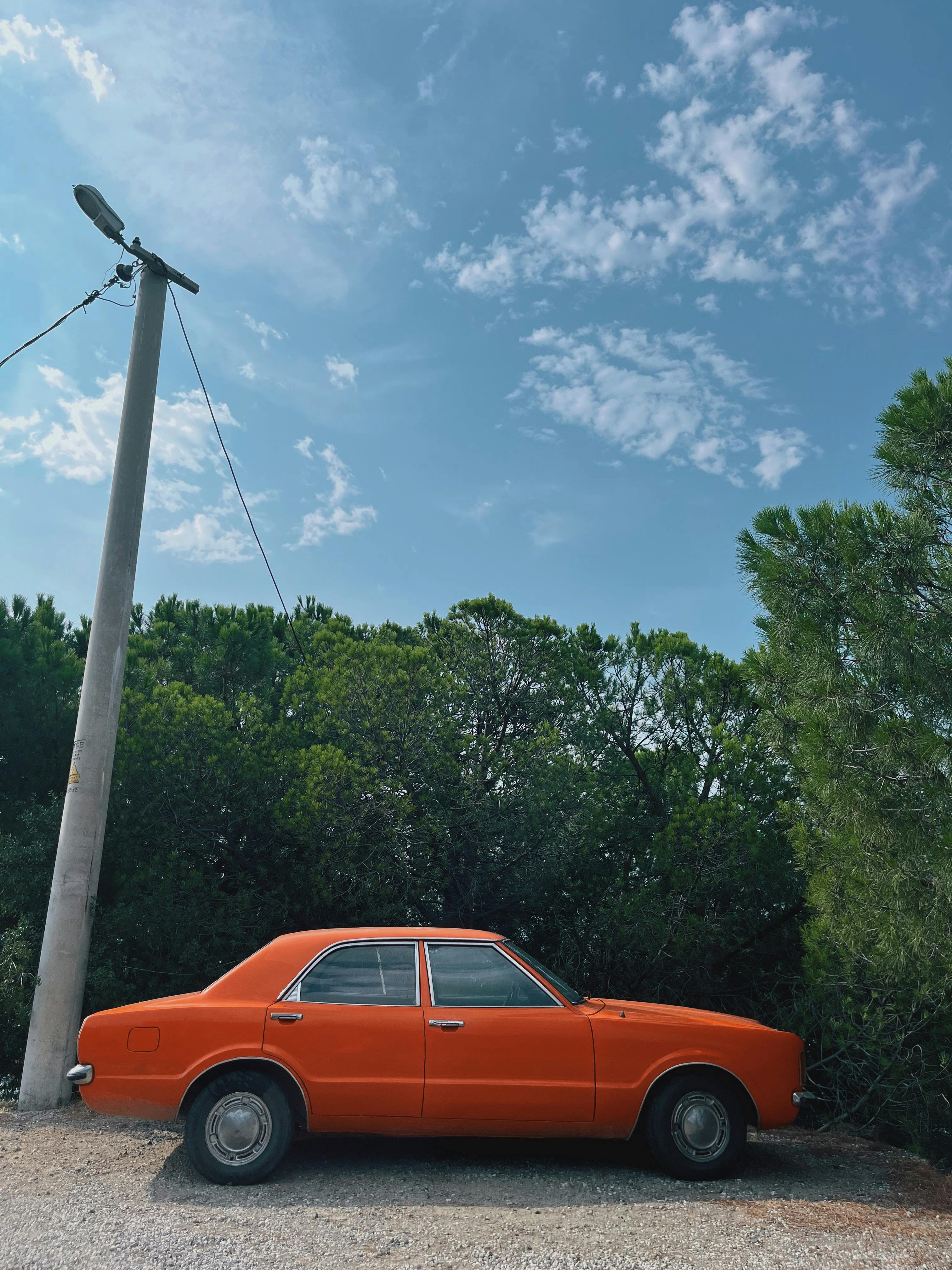 A classic orange car parked outdoors with lush green trees and a clear blue sky.
