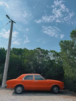 A classic orange car parked outdoors with lush green trees and a clear blue sky.