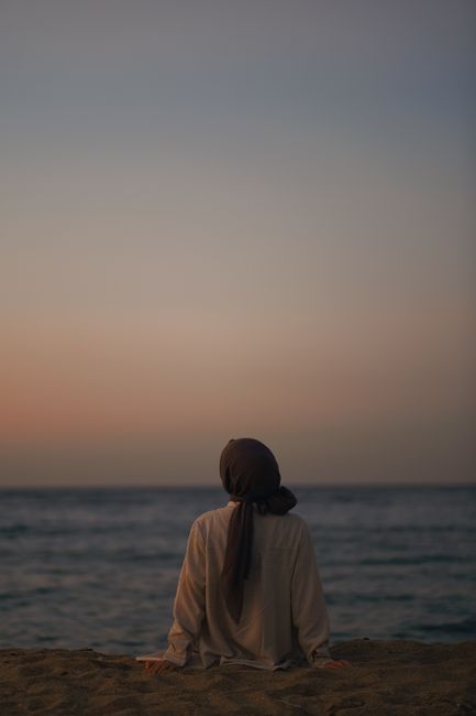 Serene scene of a person in hijab sitting by the ocean at sunset.
