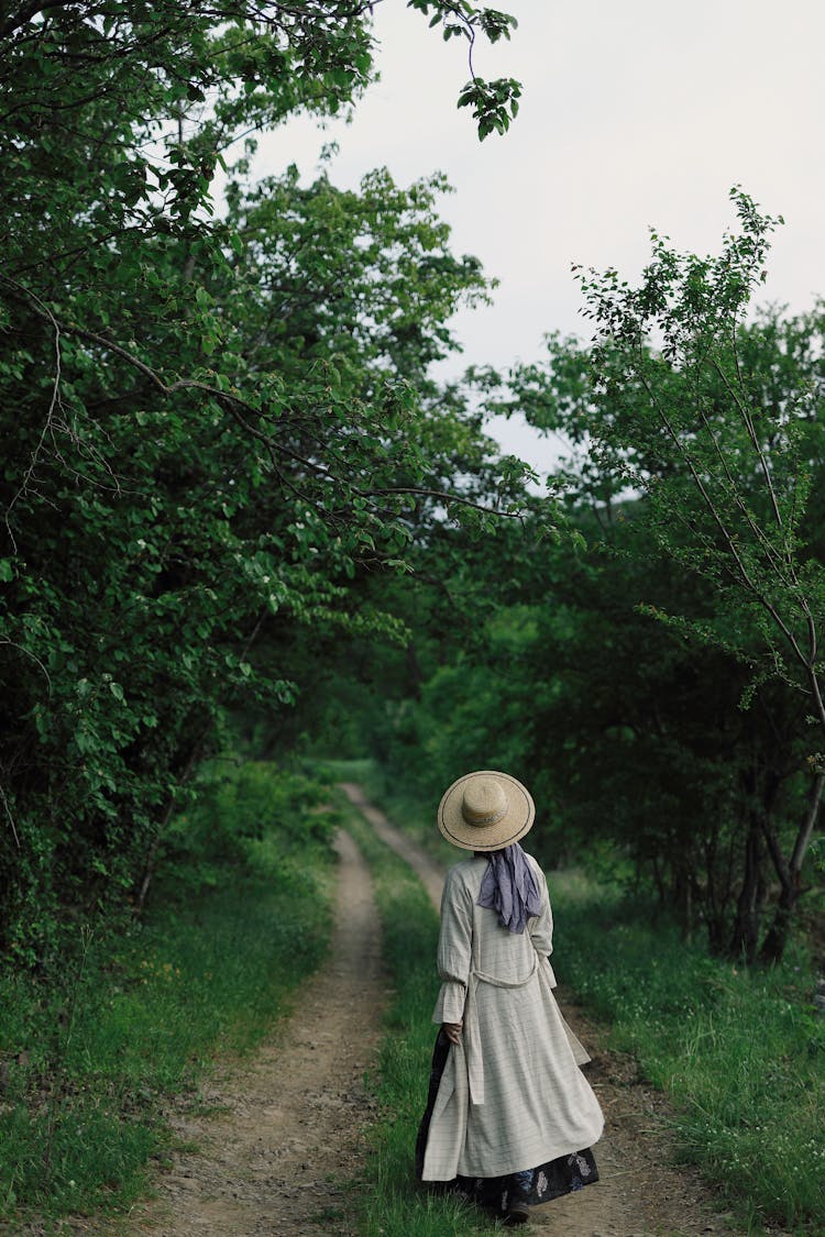 Woman In A Long Dress, Coat And A Hat Walking On A Path In Forest 