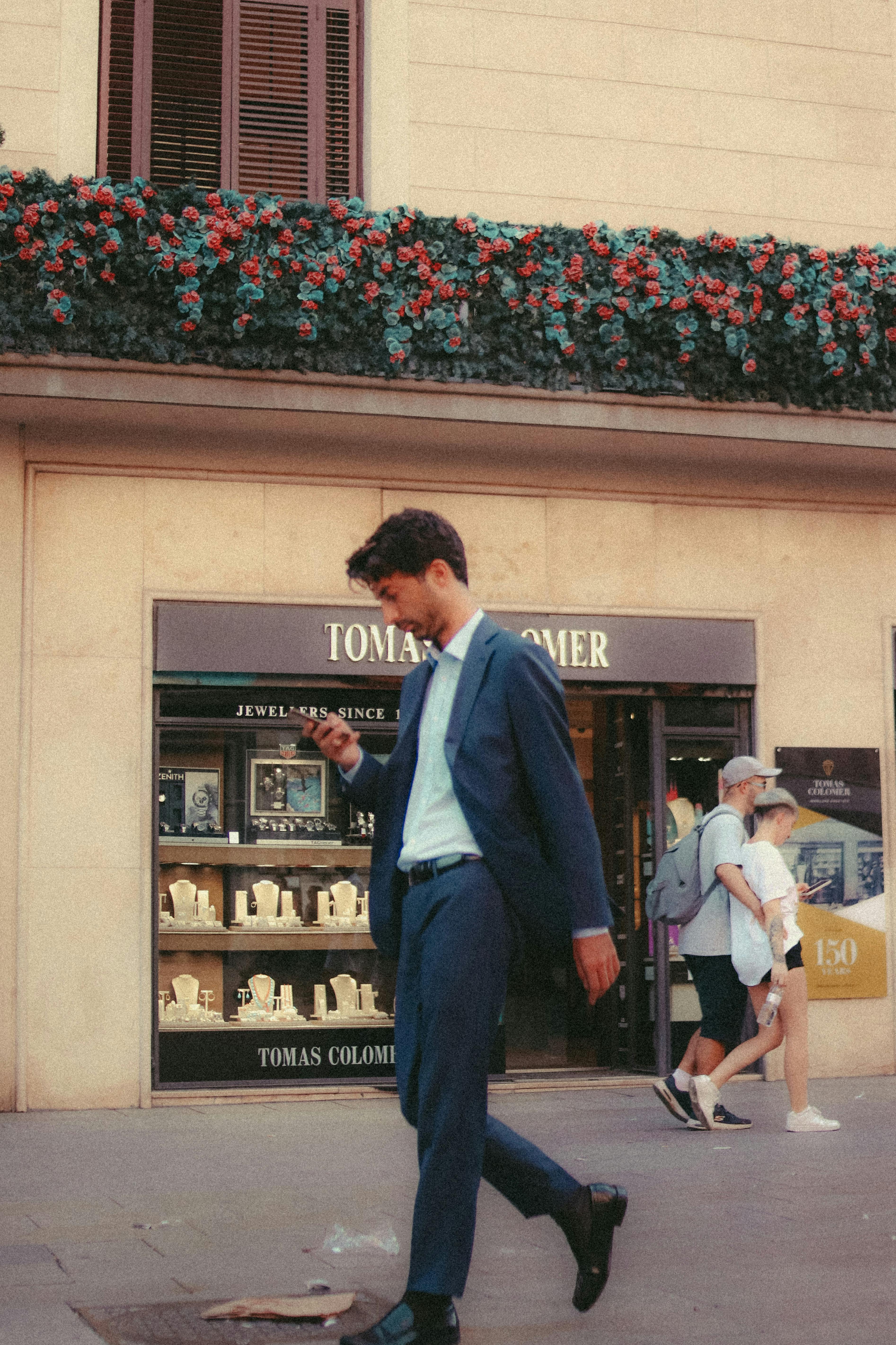 A man in a suit walks past a jewelry store, checking his phone on a city street.