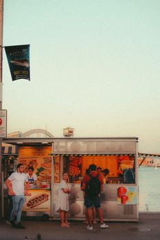 Vibrant street food stall with people enjoying snacks at sunset near a coastline. A warm, inviting scene captures urban life.