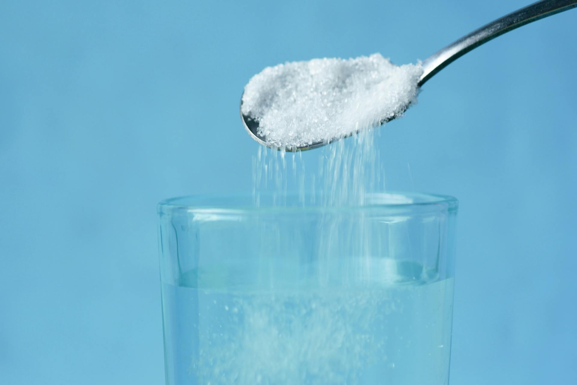 Pouring of White Sugar from Spoon in a Clear Drinking Glass with Water