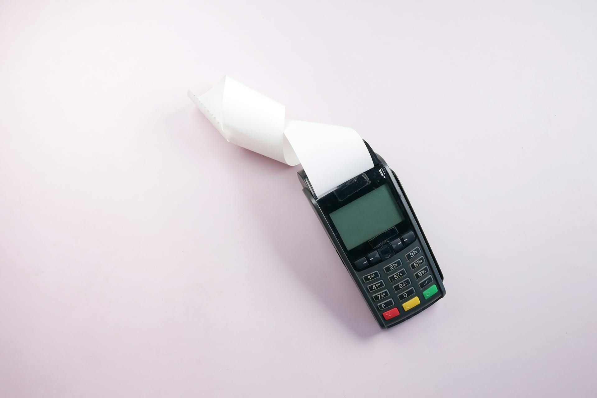 Overhead shot of a payment terminal with a receipt on a light pink background. Perfect for finance-related content.