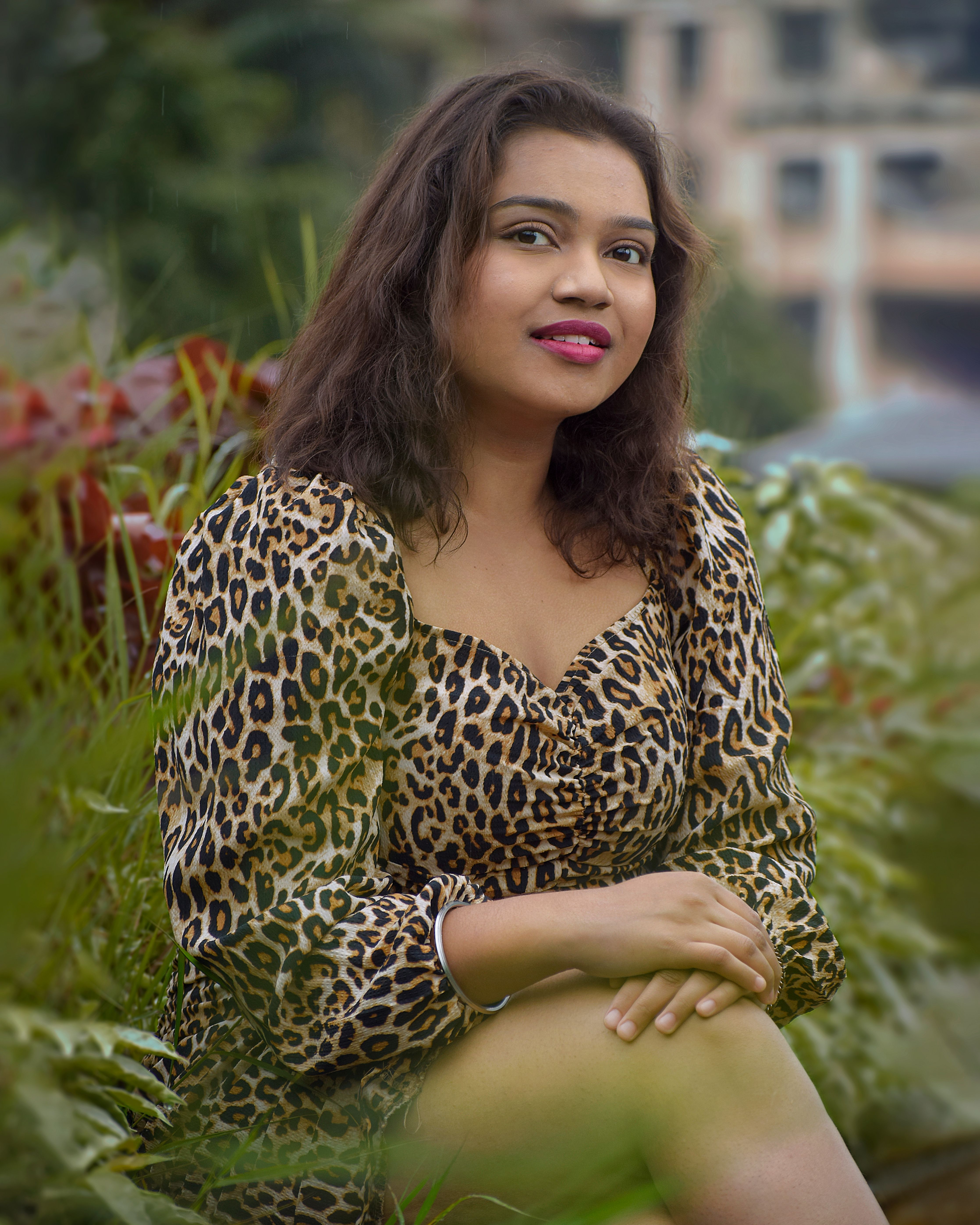 Elegant portrait of a smiling woman sitting outdoors, wearing a leopard print dress amid lush greenery.