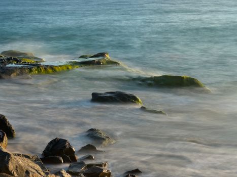 Peaceful long exposure shot of ocean water flowing over rocky shore with mossy stones.