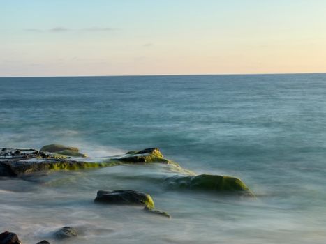 Long exposure photo of a tranquil ocean with moss-covered rocks at sunset.