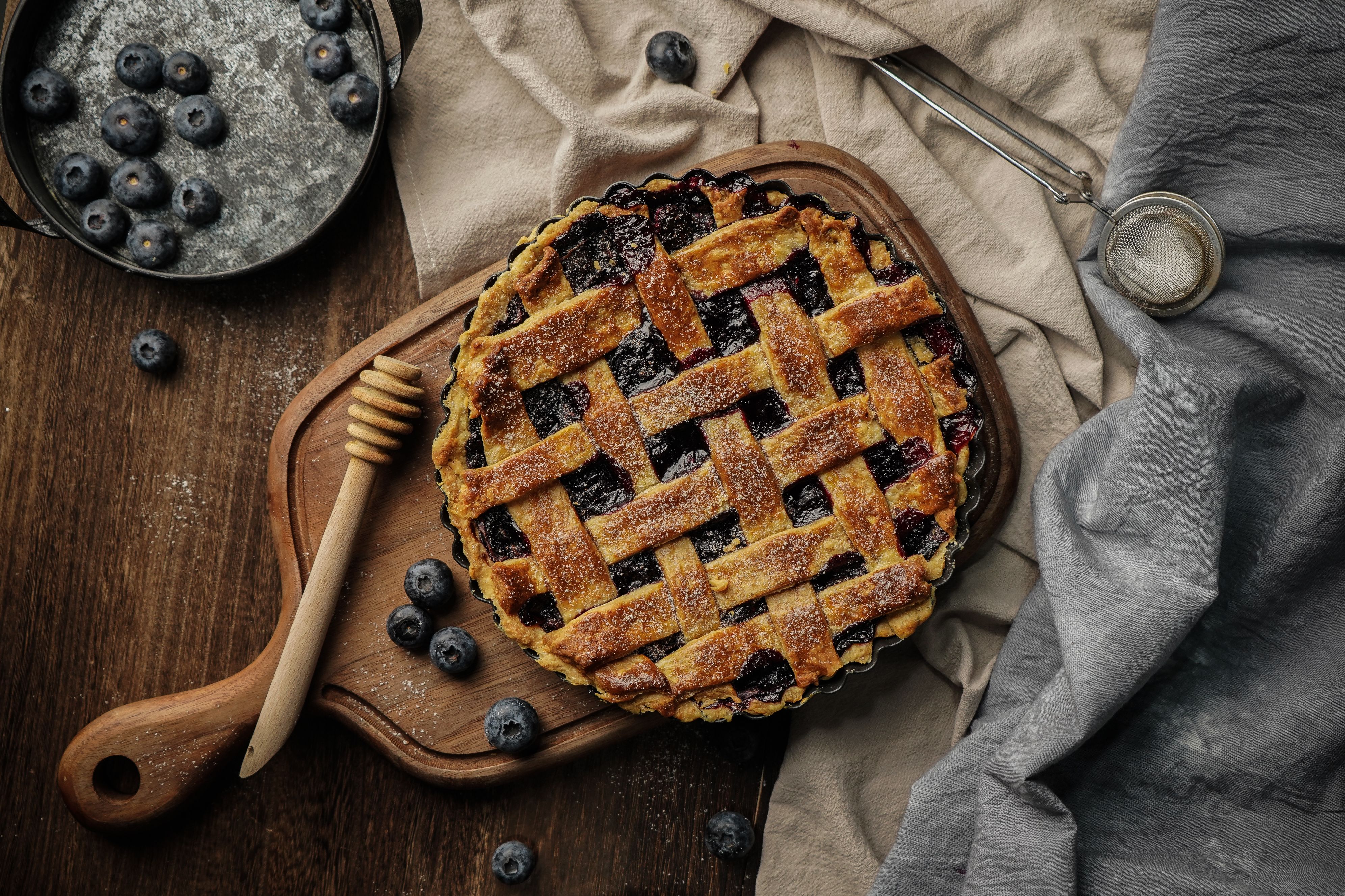 A top view of a blueberry pie with a lattice crust, surrounded by fresh blueberries on a rustic wooden board.