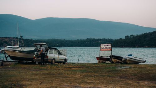 Free A man loads a car near lakeshore with boats, mountains in the background. Stock Photo