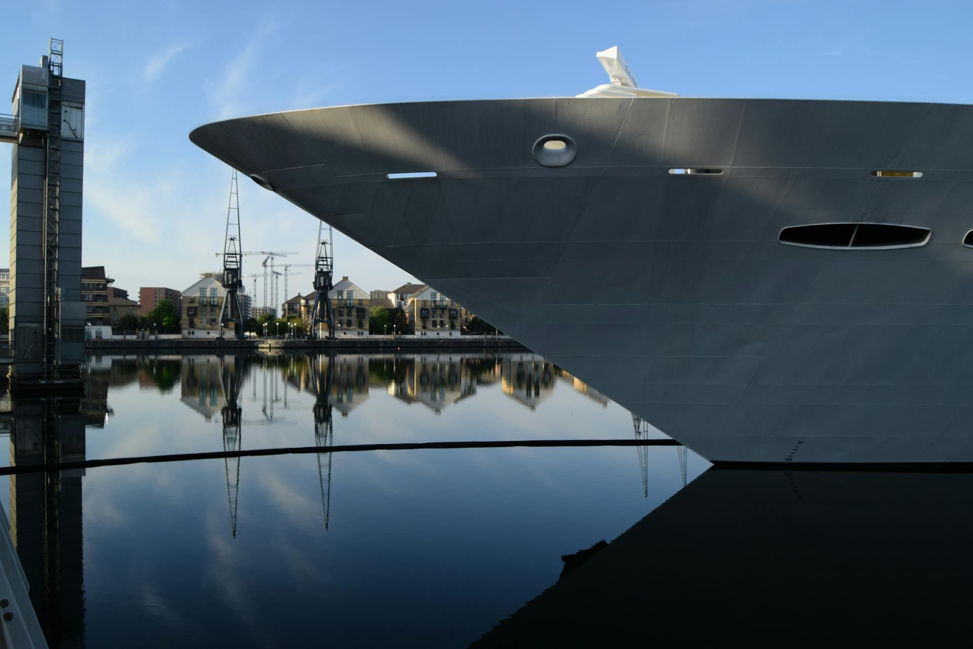 A close-up view of a ship's bow reflecting in calm waters at a dock with cranes and buildings in the background.