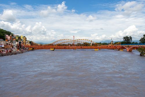 Free A vibrant rusty bridge over a bustling river with a backdrop of cloudy skies and distant hills. Stock Photo