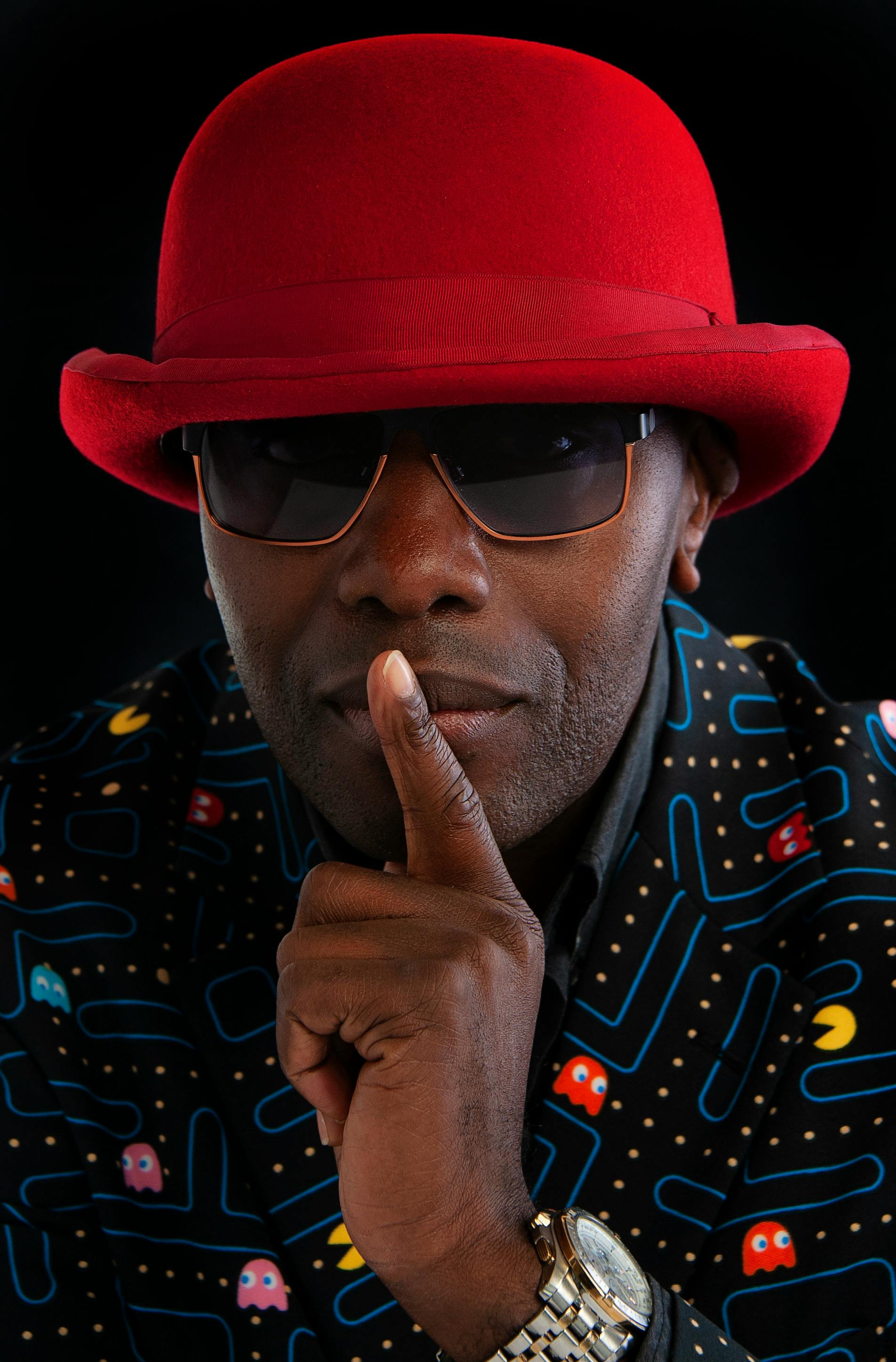 Close-up portrait of a fashionable man wearing a red hat and sunglasses, signaling silence.