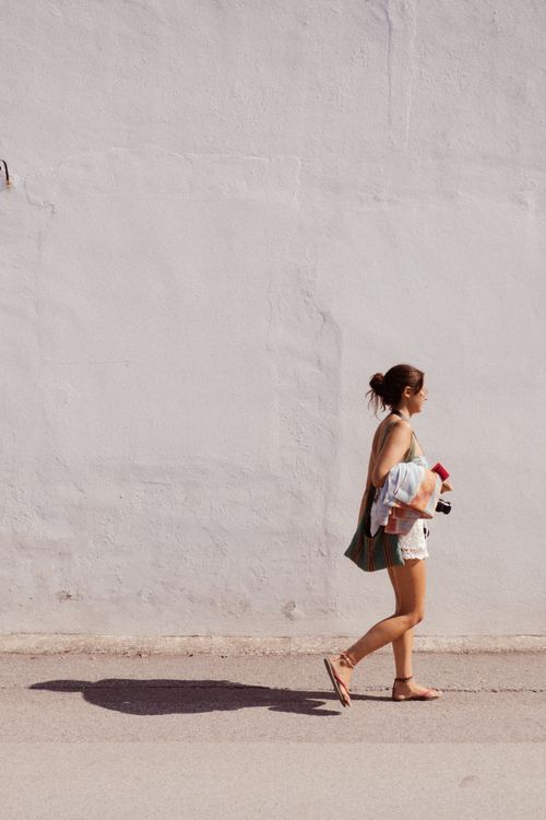 Free Sunlit scene of a woman walking past a white wall holding various items, casting a shadow. Stock Photo